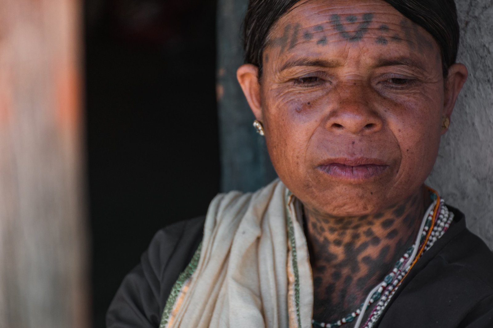 A portrait of an older woman with traditional facial and neck tattoos, wearing a dark outfit with a light-colored scarf and beaded necklaces.